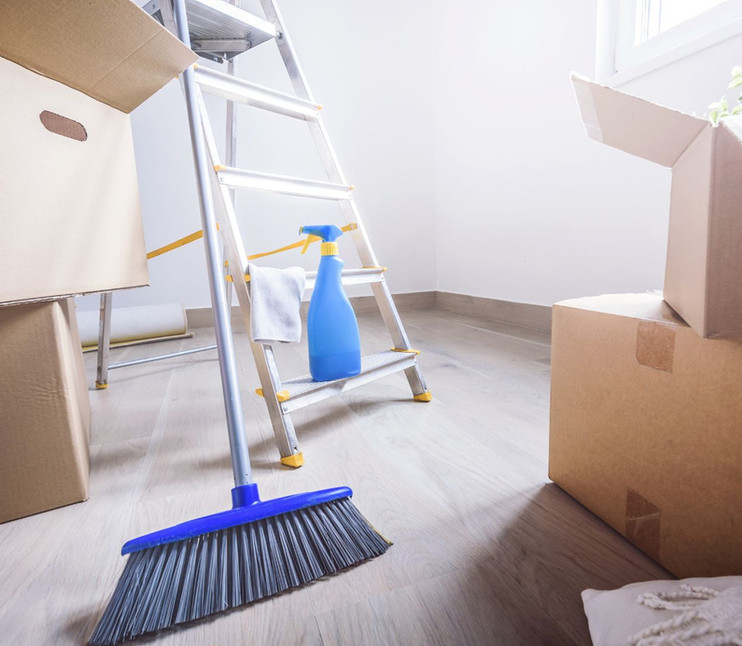 Cardboard moving boxes, blue broom, stepladder, and spray bottle in empty room.